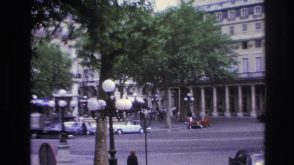 1967 Paris street scene partly obscured by a window mullion, with pedestrians, cars, globe lamps, trees, and arcaded building fronts.