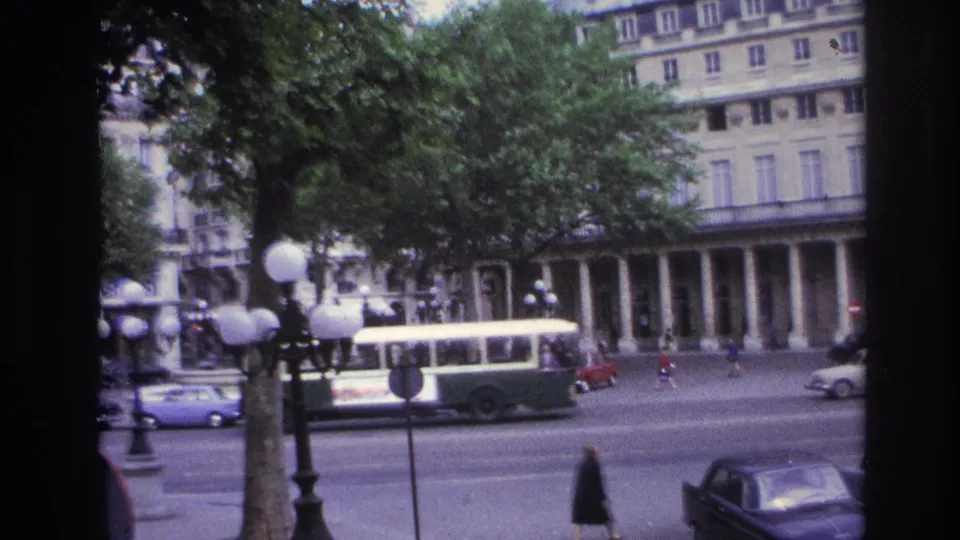 1967 Paris street footage with a green-and-white bus, classic cars, pedestrians, globe streetlamps, trees, and a foreground window frame.