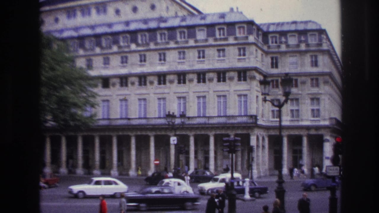 1967 Paris home-movie frame showing a grand classical building with columns, cars, pedestrians, signs, and a lamppost.