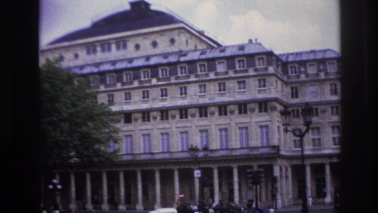 1967 Paris street view with a large classical facade, tree cover, cars, street signs, and small pedestrian figures.