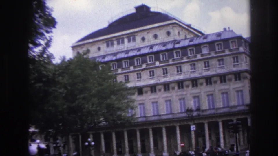 Handheld 1967 Paris frame with a tilted view of the upper facade, mansard roof, leafy trees, cars, and pedestrians below.