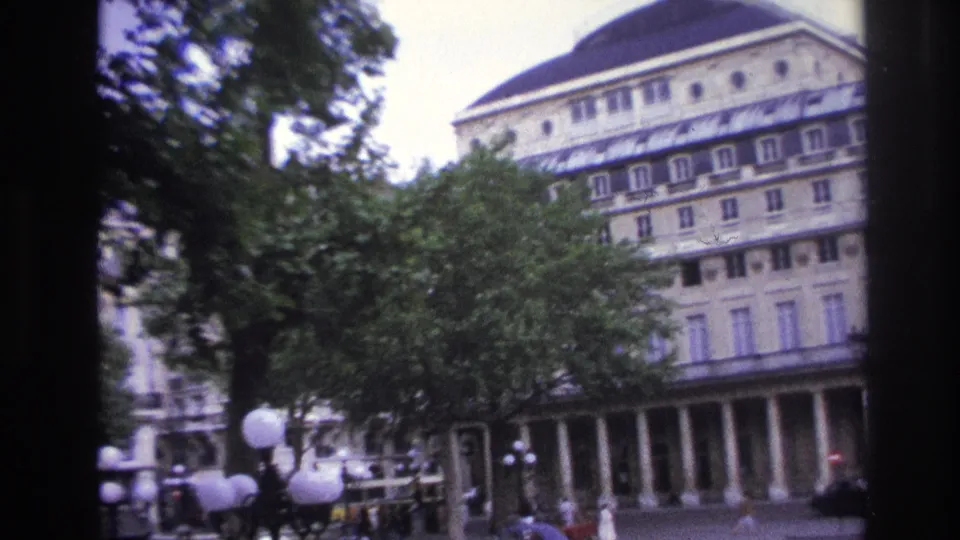 1967 Paris home-movie frame showing trees in the foreground, a columned building, street lamps, cars, and distant pedestrians.