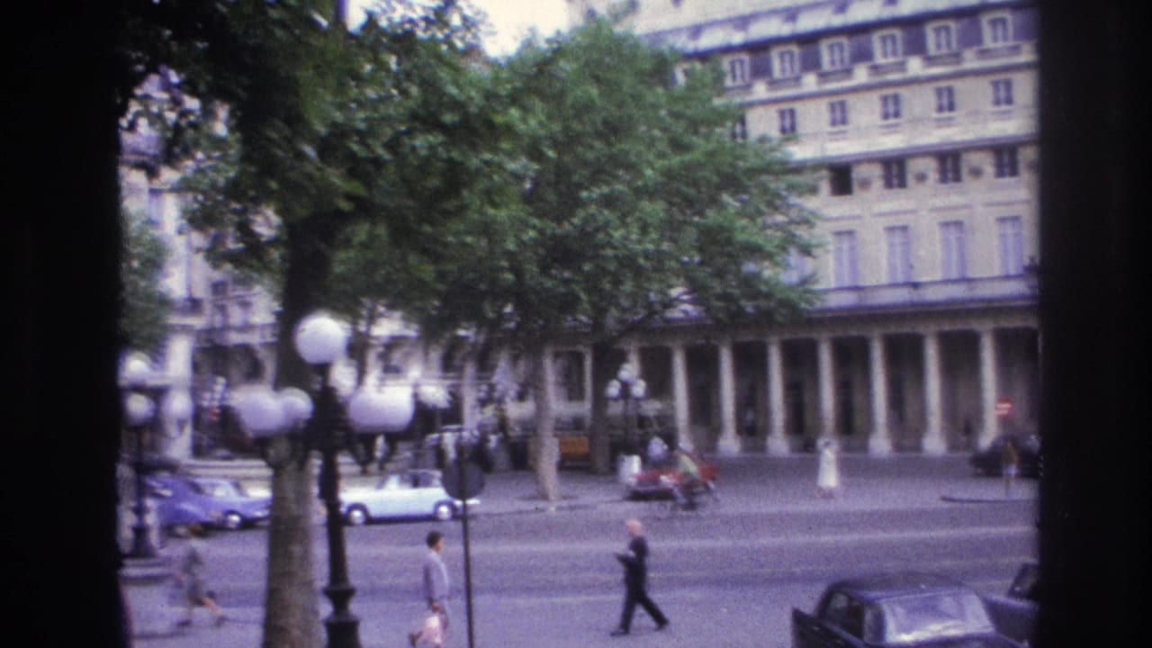 1967 Paris traffic scene with pedestrians, parked and moving cars, globe streetlamps, trees, and a large formal facade beyond the road.