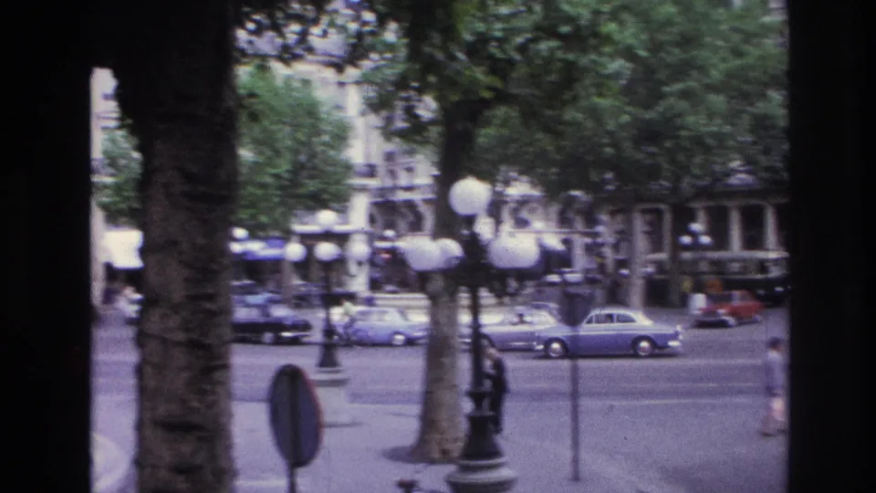 1967 Paris street view showing cars crossing the frame, globe streetlamps, tree trunks, traffic signs, and architecture beyond the roadway.