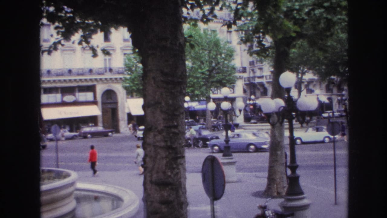 1967 Paris square viewed from behind trees and a window frame, with a fountain basin, pedestrians, cars, and globe streetlights.