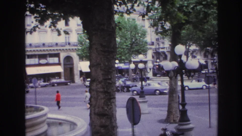 1967 Paris square viewed from behind trees and a window frame, with a fountain basin, pedestrians, cars, and globe streetlights.