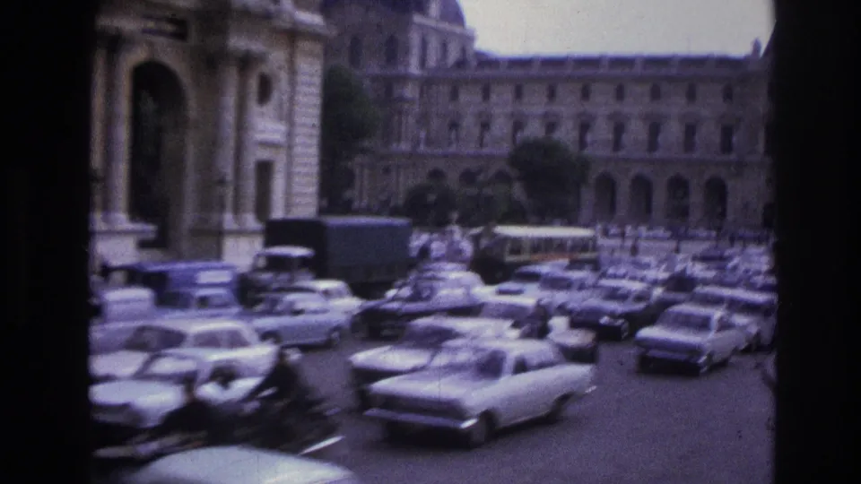 1967 Paris street scene with a line of parked cars, a bus or truck, pedestrians, stone facades, and an arched building entrance.
