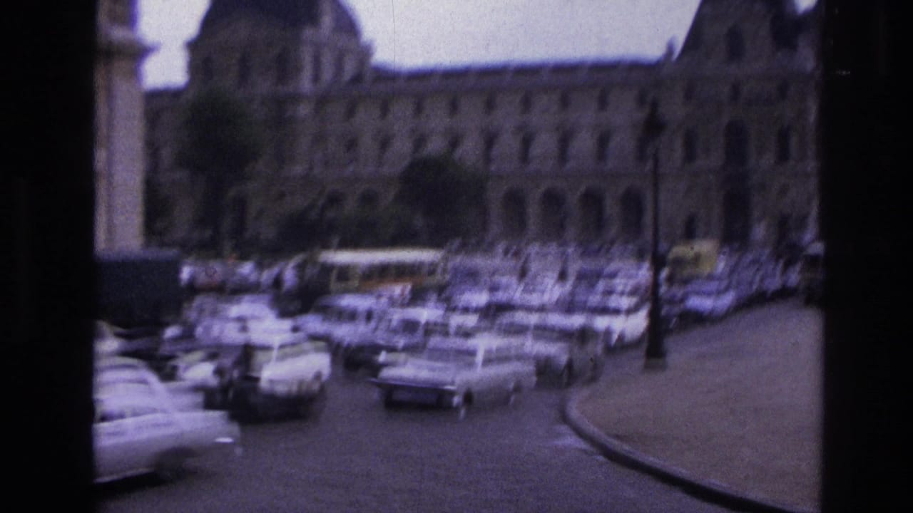 1967 Paris Super 8 frame showing the Louvre facade behind a traffic-filled street with classic cars and a soft handheld view.