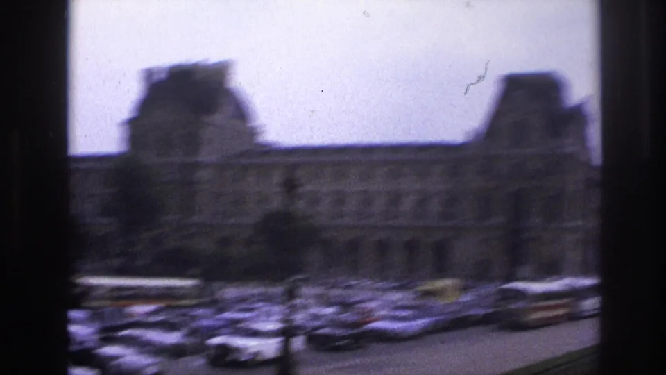 Handheld 1967 Paris view across traffic toward the Louvre, with motion blur and rows of mid-century cars.
