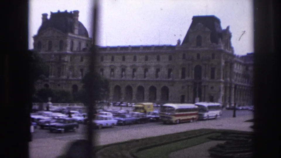 1967 Paris Super 8 frame with the Louvre exterior, rows of cars, buses, street lamps, and a garden border in the foreground.
