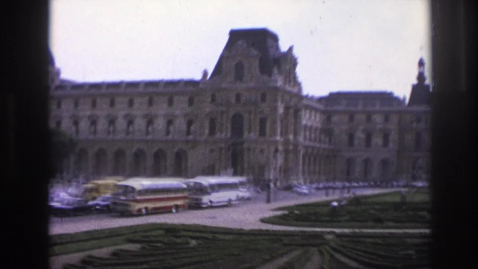 1967 Paris home-movie frame showing tour buses, cars, and clipped garden beds in front of the Louvre facade.