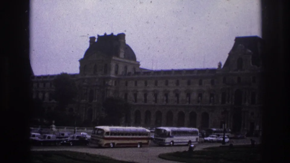 Dark-framed 1967 Paris home-movie view of the Louvre exterior, with tour buses, parked cars, and small pedestrians near the grounds.
