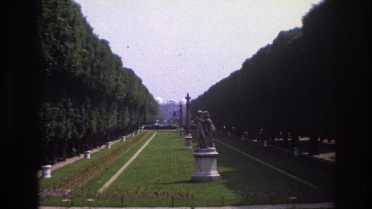 1967 Paris home-movie view of a formal garden with clipped hedges, a central lawn, lampposts, flowerbeds, and a stone statue.