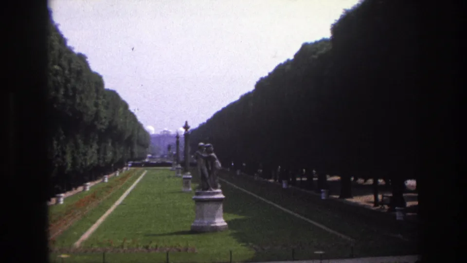 1967 Paris travel footage frames a formal garden aisle with dense clipped hedges, a green lawn, lampposts, and a classical statue.