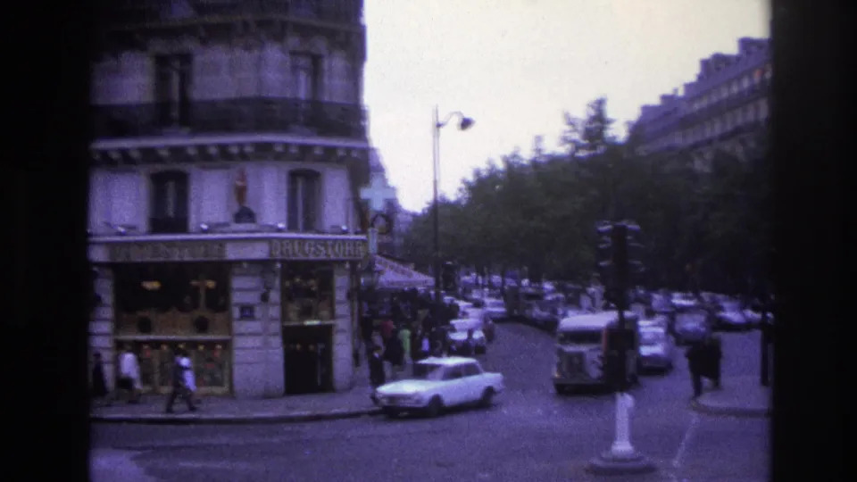 1967 Paris street frame with a white sedan near a drugstore corner, pedestrians, parked cars, and a traffic signal.