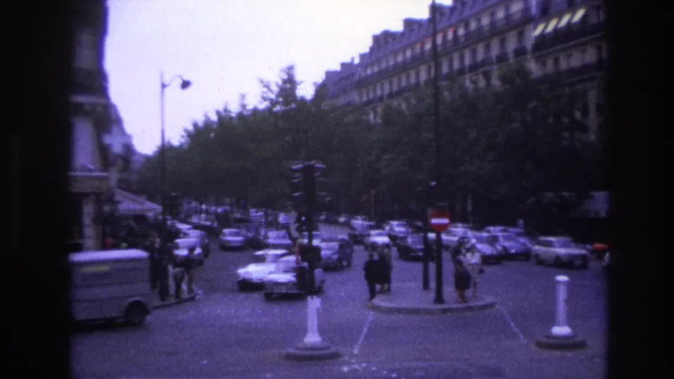 1967 Paris home-movie frame looking down a busy tree-lined street with cars, pedestrians, bollards, and apartment facades.