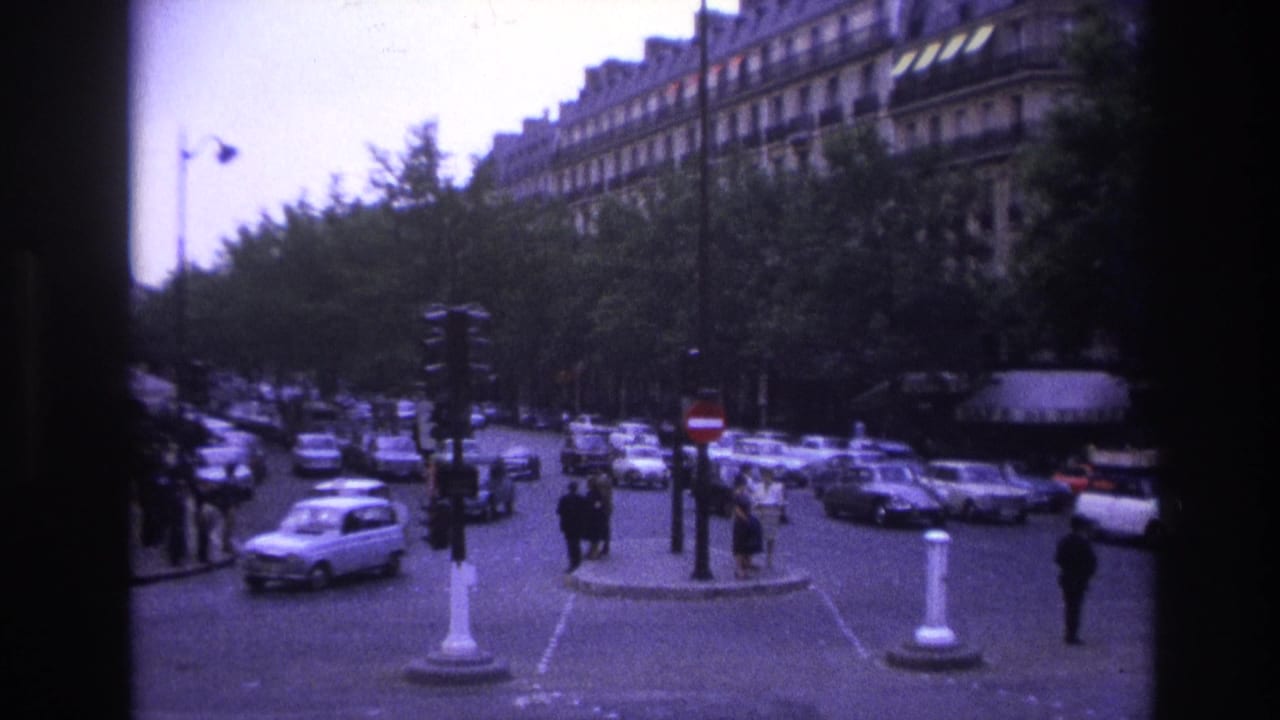 Busy Paris intersection with vintage cars, pedestrians, trees, and apartment facades.