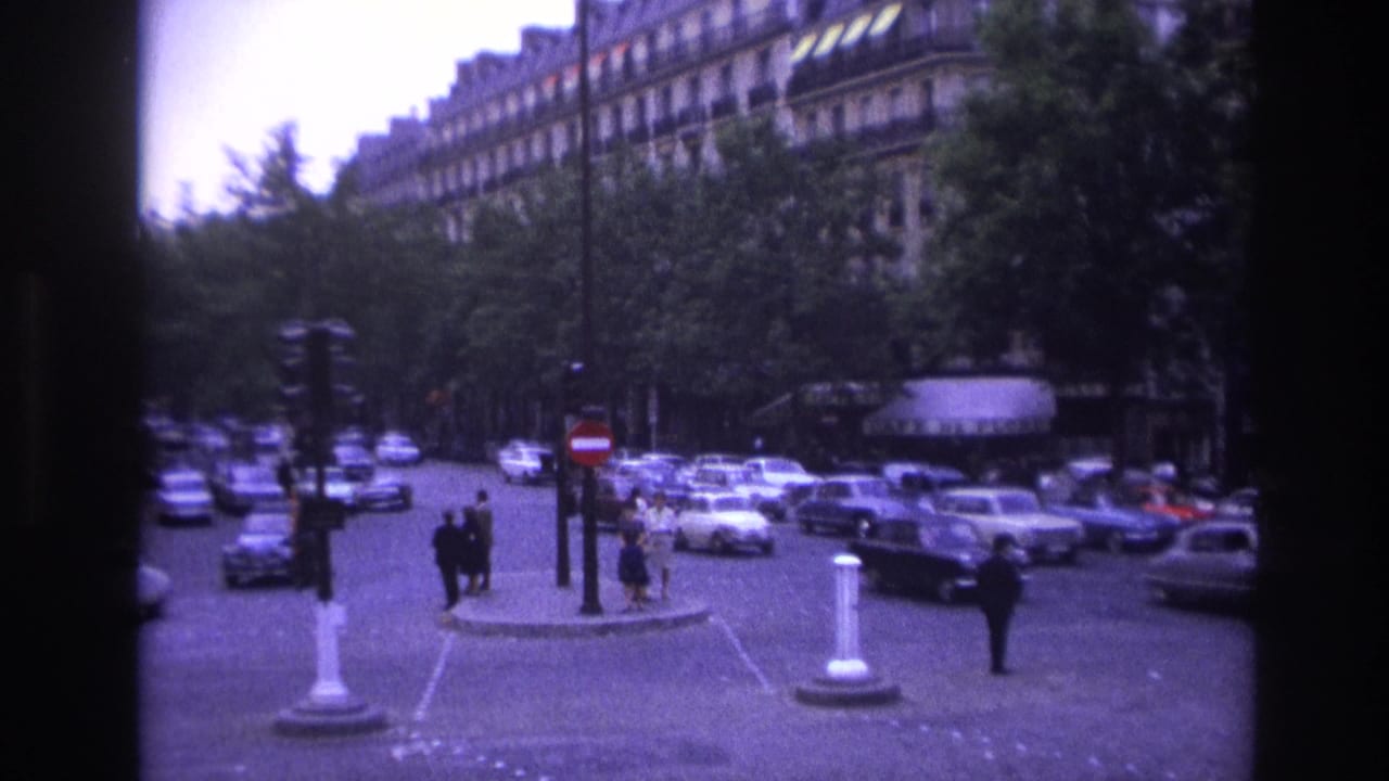 1967 Paris intersection with white and dark sedans, pedestrians on a small island, and Haussmann-style buildings behind trees.