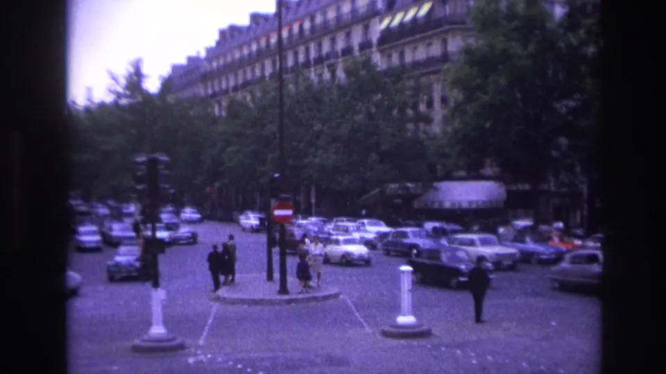 1967 Paris intersection with white and dark sedans, pedestrians on a small island, and Haussmann-style buildings behind trees.