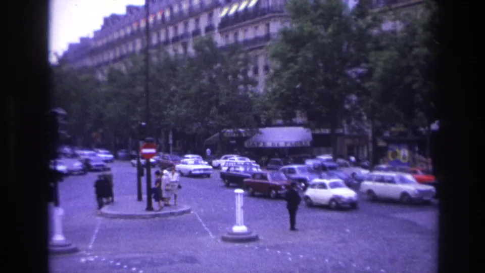 1967 Paris street scene showing period automobiles, pedestrians, bollards, trees, and shop awnings along a busy intersection.