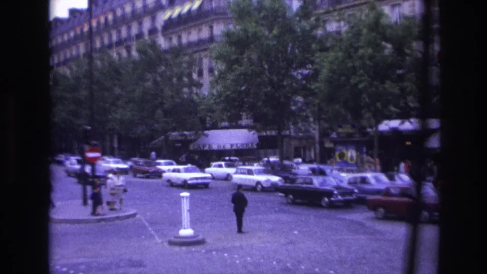 1967 Paris intersection framed by dark film edges, with period traffic, pedestrians, white bollards, trees, and apartment facades.