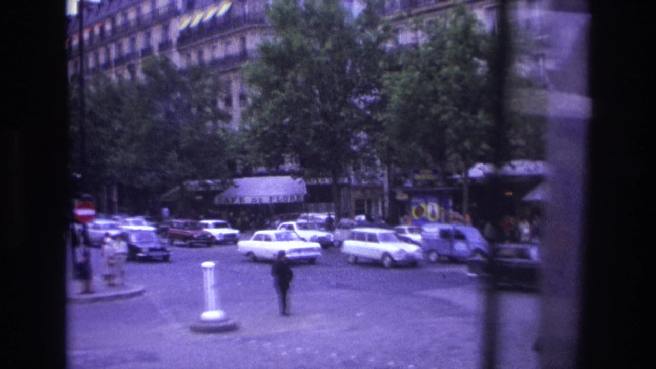 1967 Paris street intersection with white sedans, leafy trees, ornate buildings, and a lone pedestrian in handheld 8mm footage.