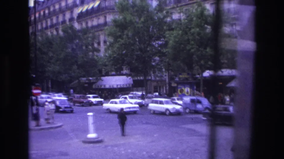 1967 Paris street intersection with white sedans, leafy trees, ornate buildings, and a lone pedestrian in handheld 8mm footage.