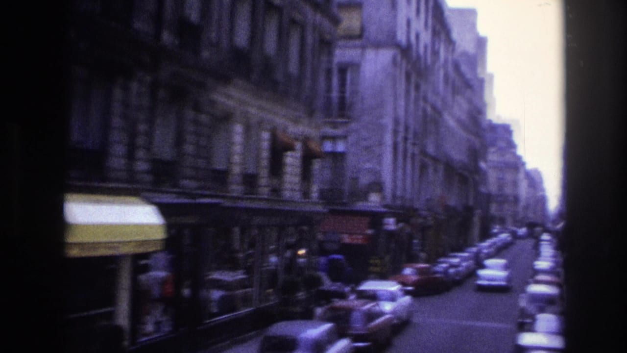1967 Paris side street with curbside cars, yellow and red shop awnings, tall stone facades, and distant pedestrians.