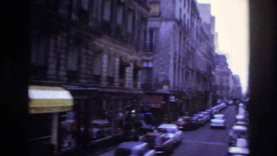 1967 Paris side street with curbside cars, yellow and red shop awnings, tall stone facades, and distant pedestrians.