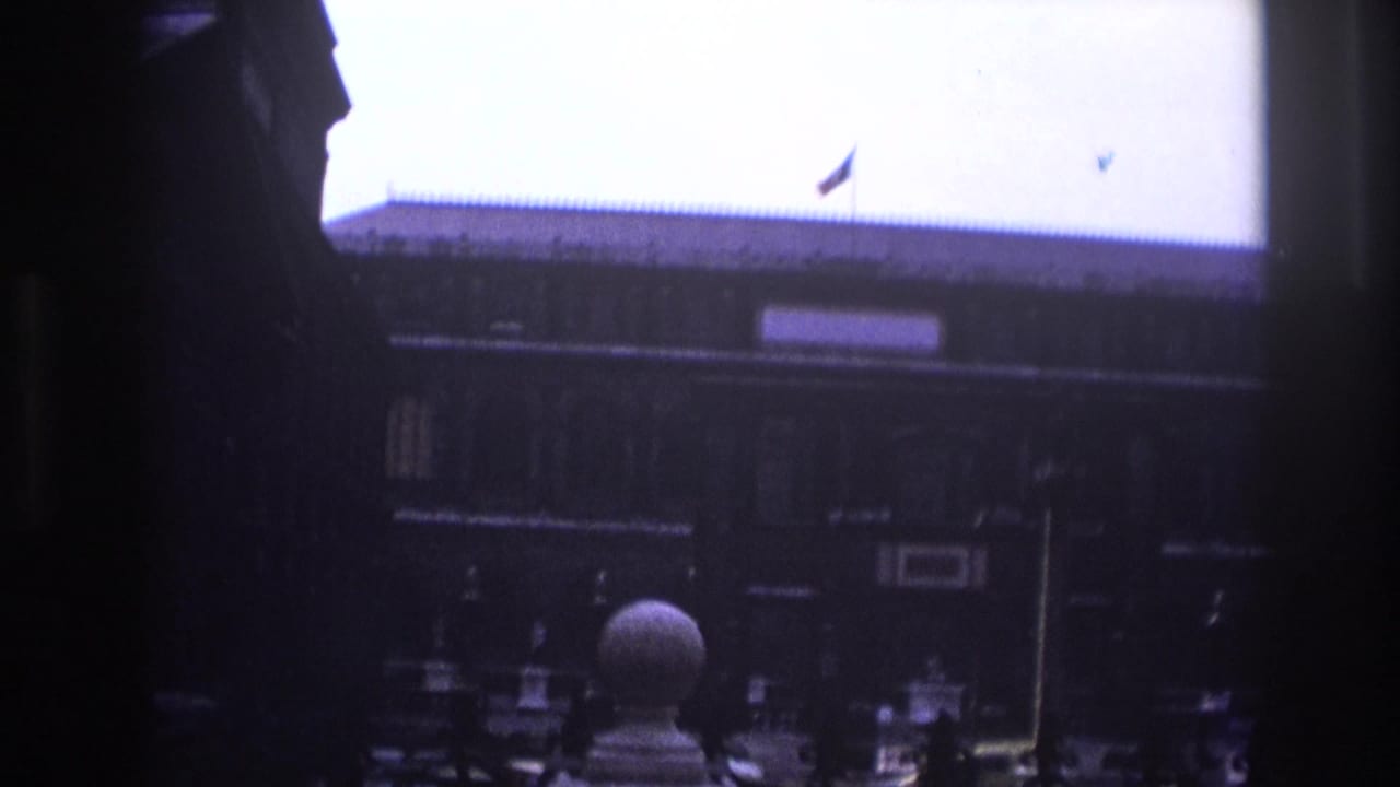 1967 Paris home-movie frame showing a dark foreground edge around a historic courtyard facade with a roof flag and stone ornament.