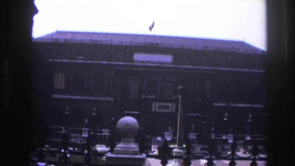 1967 Paris courtyard view framed by shadow, showing a long dark building, iron railings, a roof flag, and stone decorative forms.