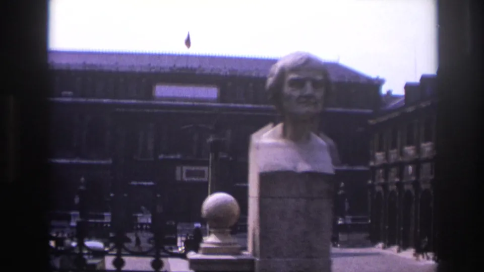 1967 Paris home-movie frame with a stone bust in the foreground, a dark courtyard facade behind it, and ironwork along the right side.