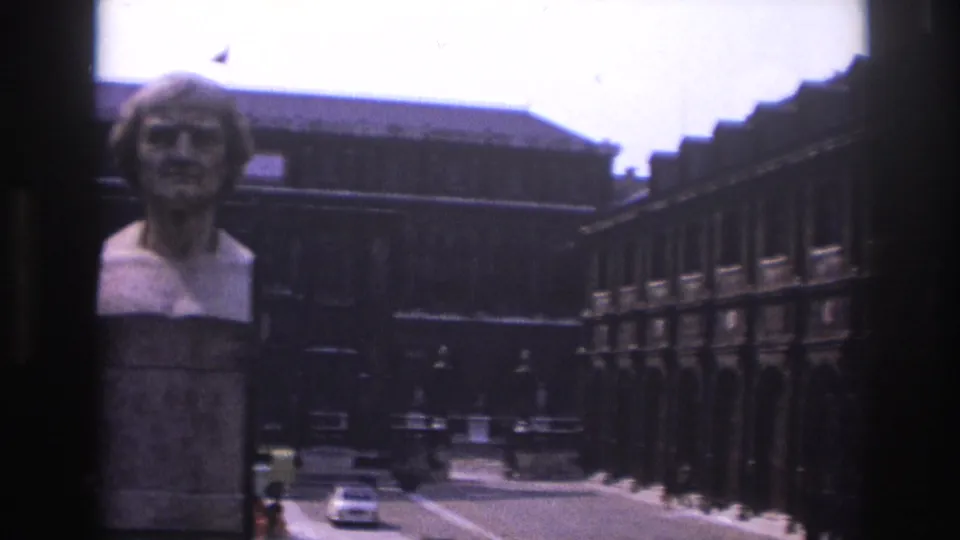 1967 Paris courtyard view with a stone bust in front, a white car and small pedestrians in the distance, and ironwork along the right side.