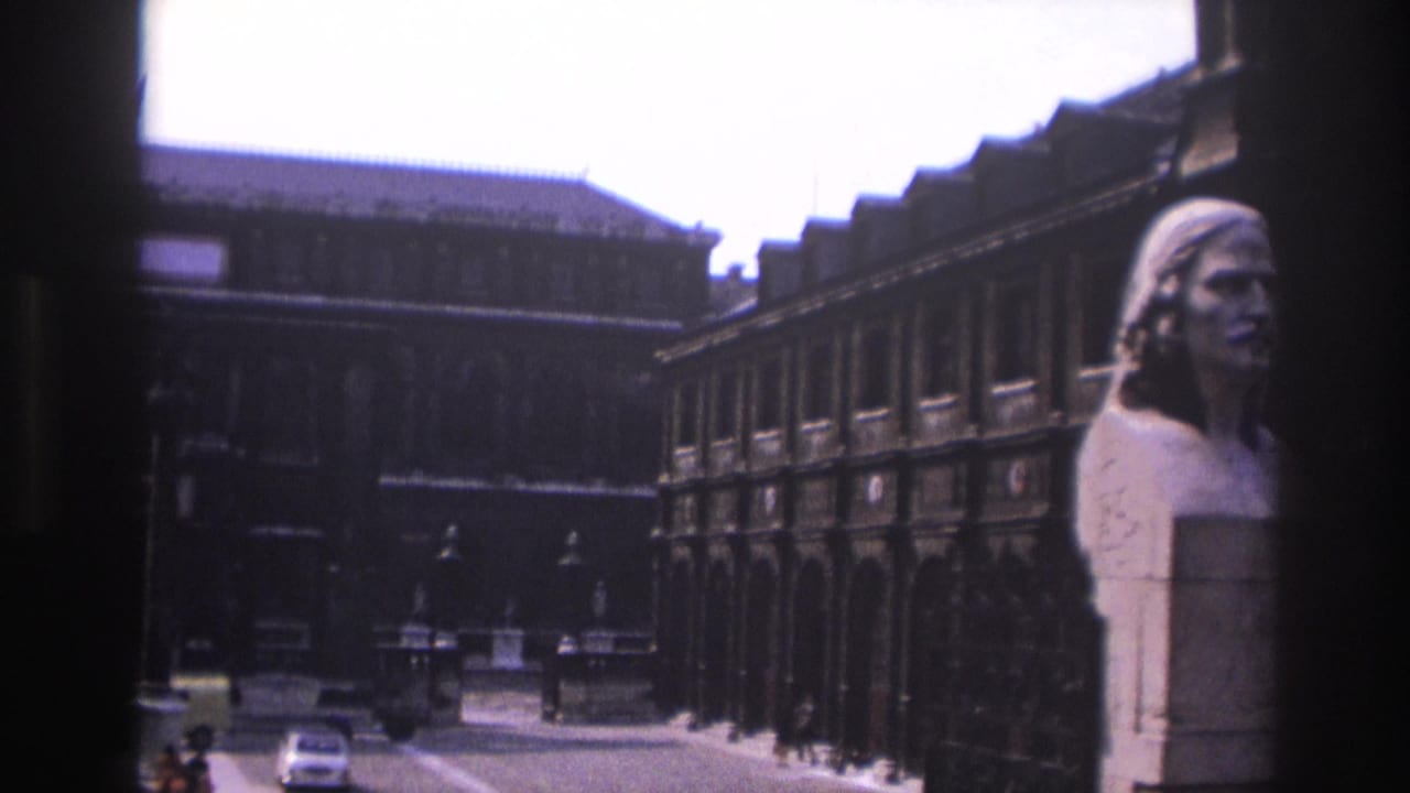 1967 8mm view of a Paris courtyard or street edge with a marble bust, ornate stone architecture, iron railing, and distant car.
