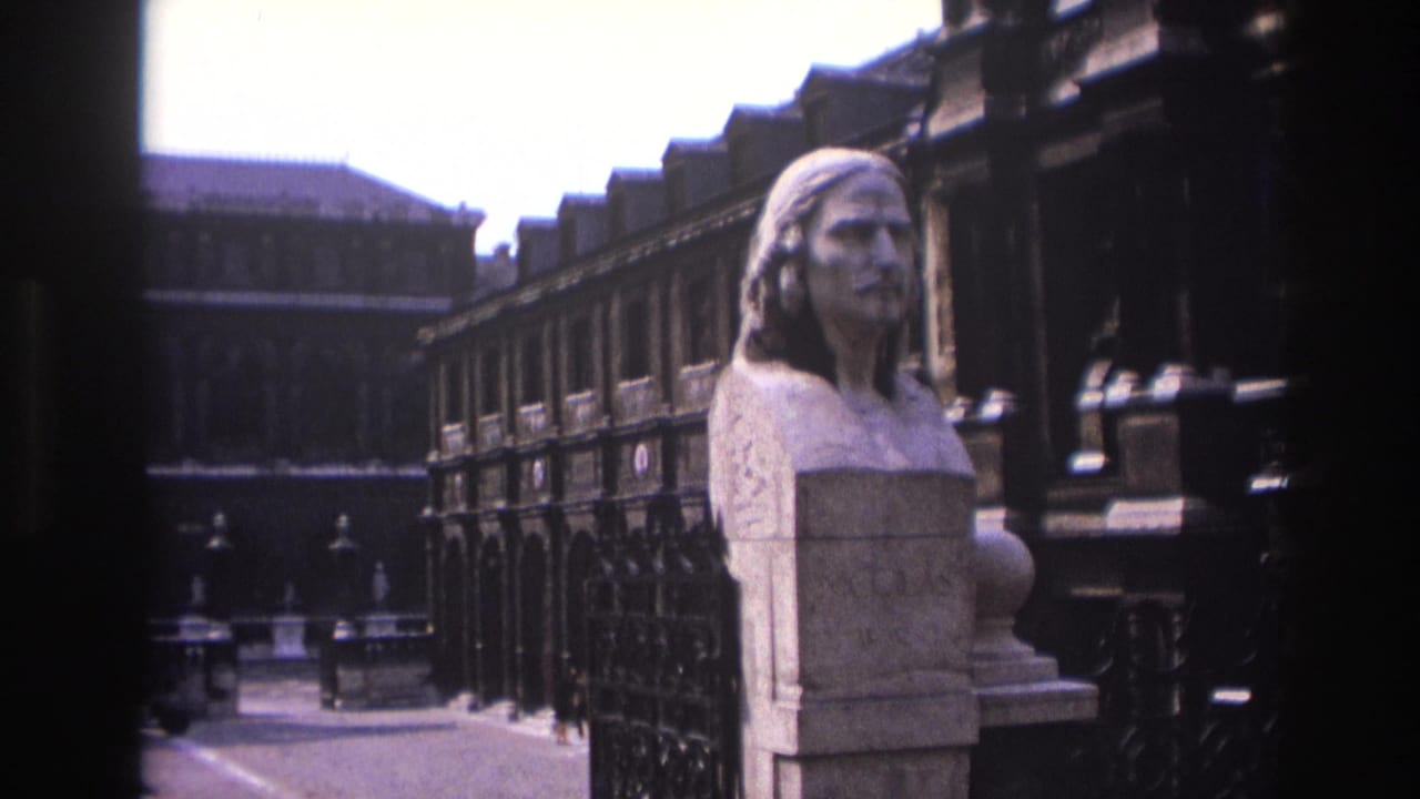 1967 Paris home-movie frame showing a marble bust close to the camera, with ironwork and historic stone facades behind it.