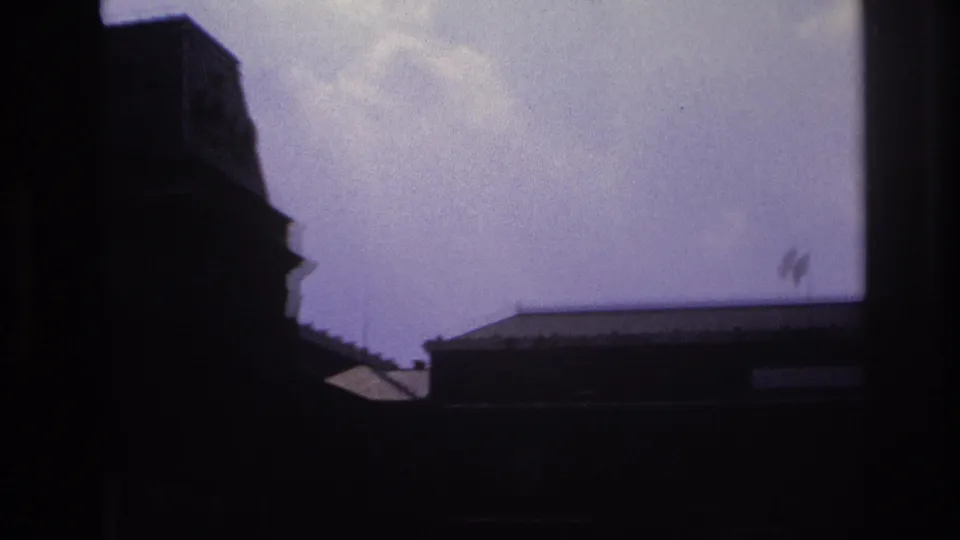 1967 Paris home-movie frame showing a dark building edge below open sky, with small flags visible near the right side.