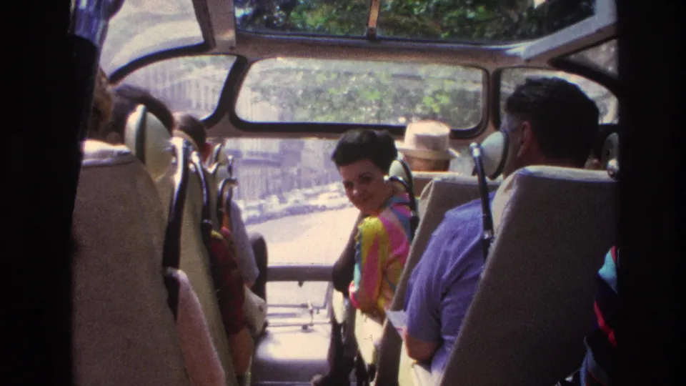 1967 Paris bus-tour passengers sit under open windows, including a woman in a colorful striped top looking back.