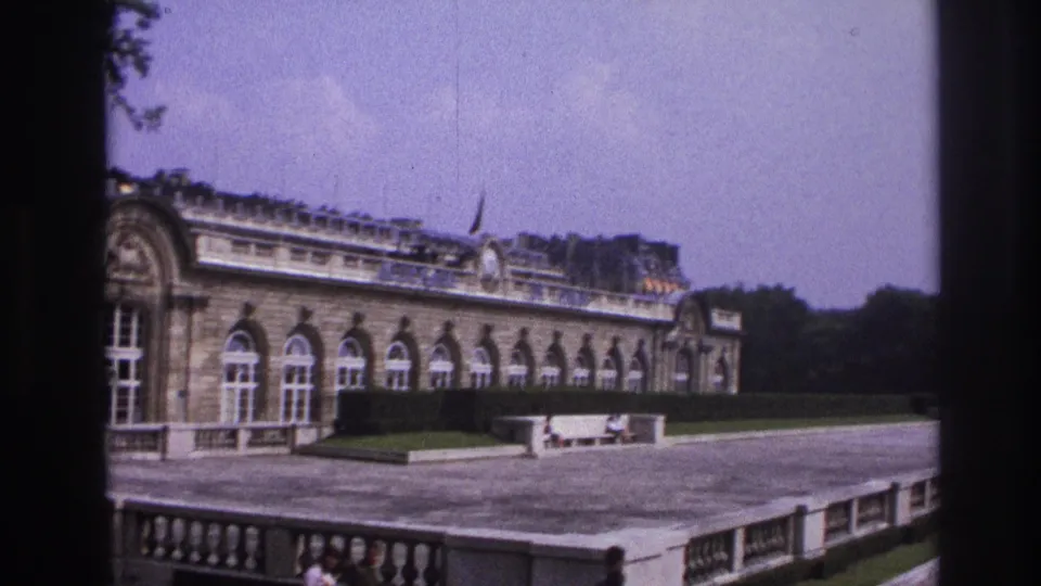 1967 Paris travel footage showing an ornate long building with arched windows, terrace paving, hedges, and small visitors below.