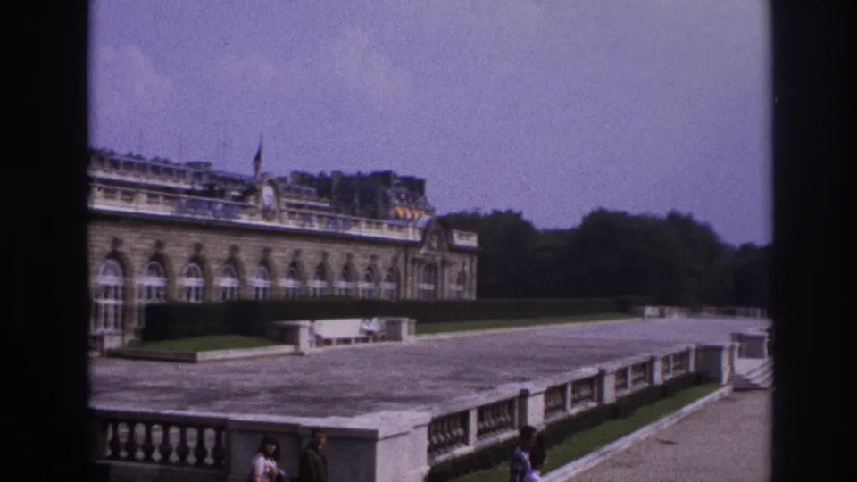Handheld 8mm Paris view of an ornate facade, terrace balustrade, clipped lawns, trees, and distant visitors on the grounds.