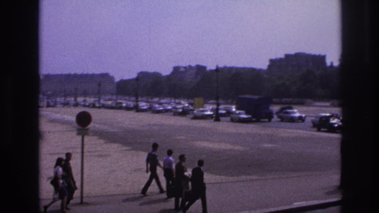 Paris street scene from 1967 showing pedestrians in period clothing near a wide road, parked cars, trees, and distant buildings.