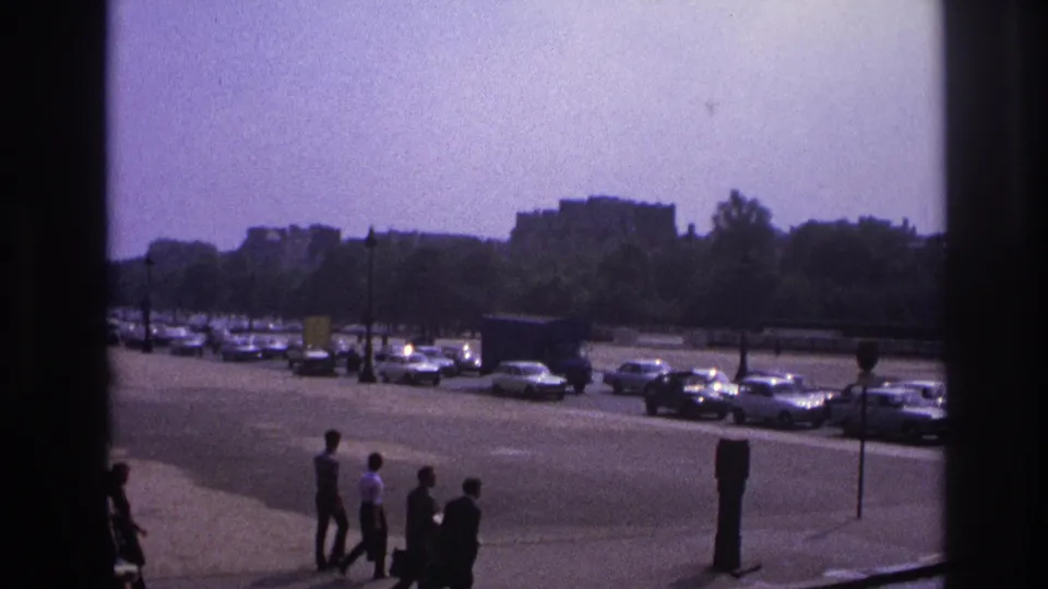 1967 Paris home-movie frame with pedestrians on a sidewalk, a broad traffic area, parked 1960s cars, and trees beyond the road.