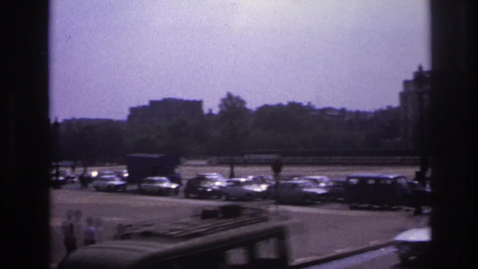 Paris street view from 1967 with a passing vehicle in the foreground, pedestrians, parked cars, trees, and distant urban buildings.