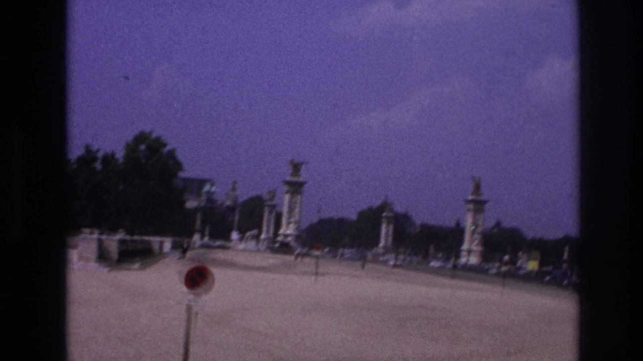 1967 Paris home-movie frame showing a vehicle approaching a broad Seine bridge with stone pylons, distant cars, trees, and pedestrians.