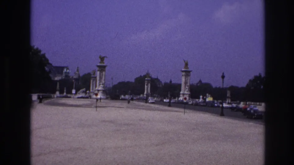 1967 Paris frame looking along a wide bridge approach with decorative pylons, small pedestrians, traffic, and a purple-toned sky.