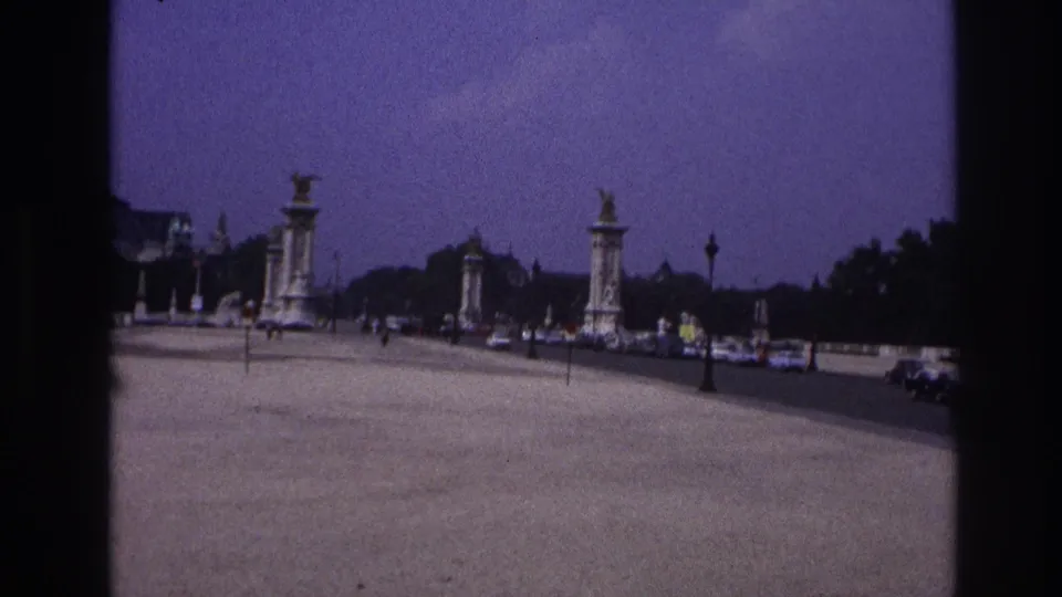 1967 Paris home-movie still showing the bridge deck from a moving viewpoint, with cars, pedestrians, pylons, and tree-lined edges.