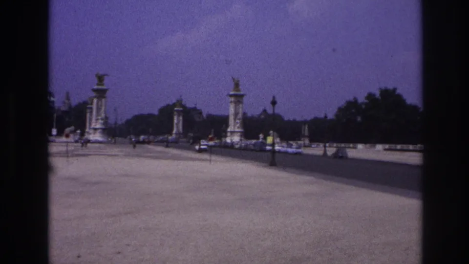 Final 1967 Paris frame showing a broad bridge approach with tall sculptural pylons, distant traffic, pedestrians, trees, and soft 8mm color.