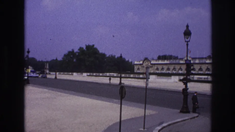 1967 Paris home-movie frame showing riverside architecture, a decorative streetlamp, signposts, cars, and distant pedestrians.