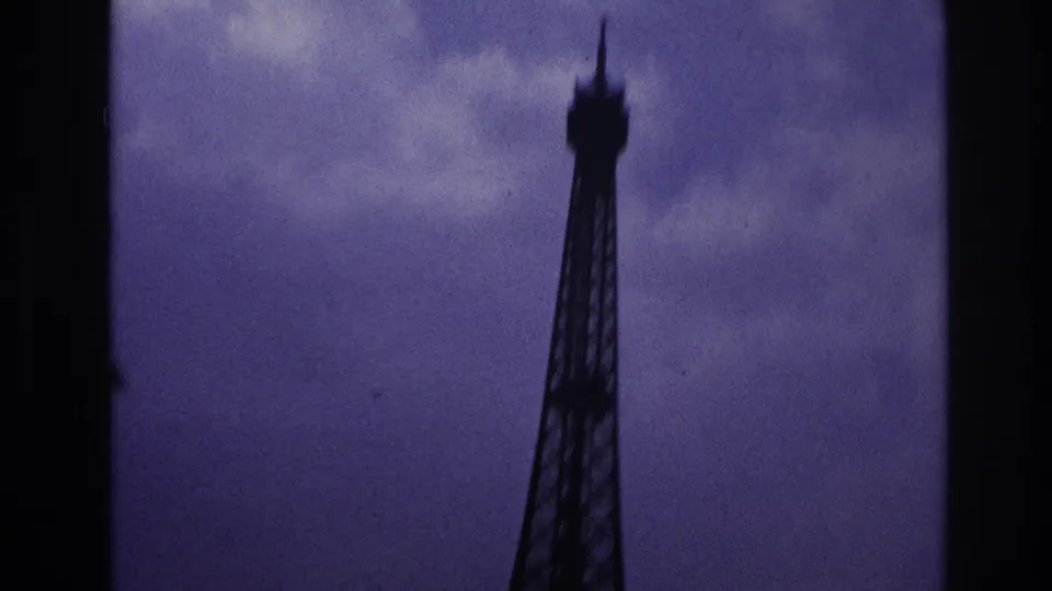 1967 Paris home-movie frame showing the upper Eiffel Tower in silhouette beneath a cloudy, purple-shifted sky.