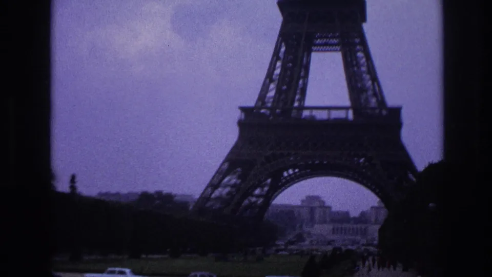 1967 Paris view of the lower Eiffel Tower with Champ de Mars lawns, distant buildings, cars, and small pedestrians.