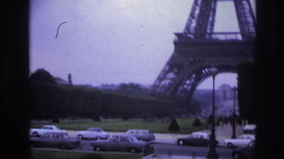 1967 Paris home-movie frame showing the Eiffel Tower base, roadway traffic, green lawns, and distant pedestrians.